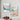 Two framed artworks of colorful cityscapes on a wall with a bowl of plums below.