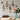 The image shows a wooden desk topped with a vase filled with a pink rose next to a wall adorned with four 
ornate vintage style picture frames with vintage art print in them and two swallow wall decor.