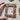 The image shows a hand holding a small, ornate gold picture frame with a painting of red leaves on a white background. The frame is intricately designed and has a vintage or antique look to it. The painting inside the frame depicts a few branches with red leaves against a white background. The leaves are painted in a delicate, watercolor style, and the branches are thin and curved. The overall effect is one of simplicity and elegance.